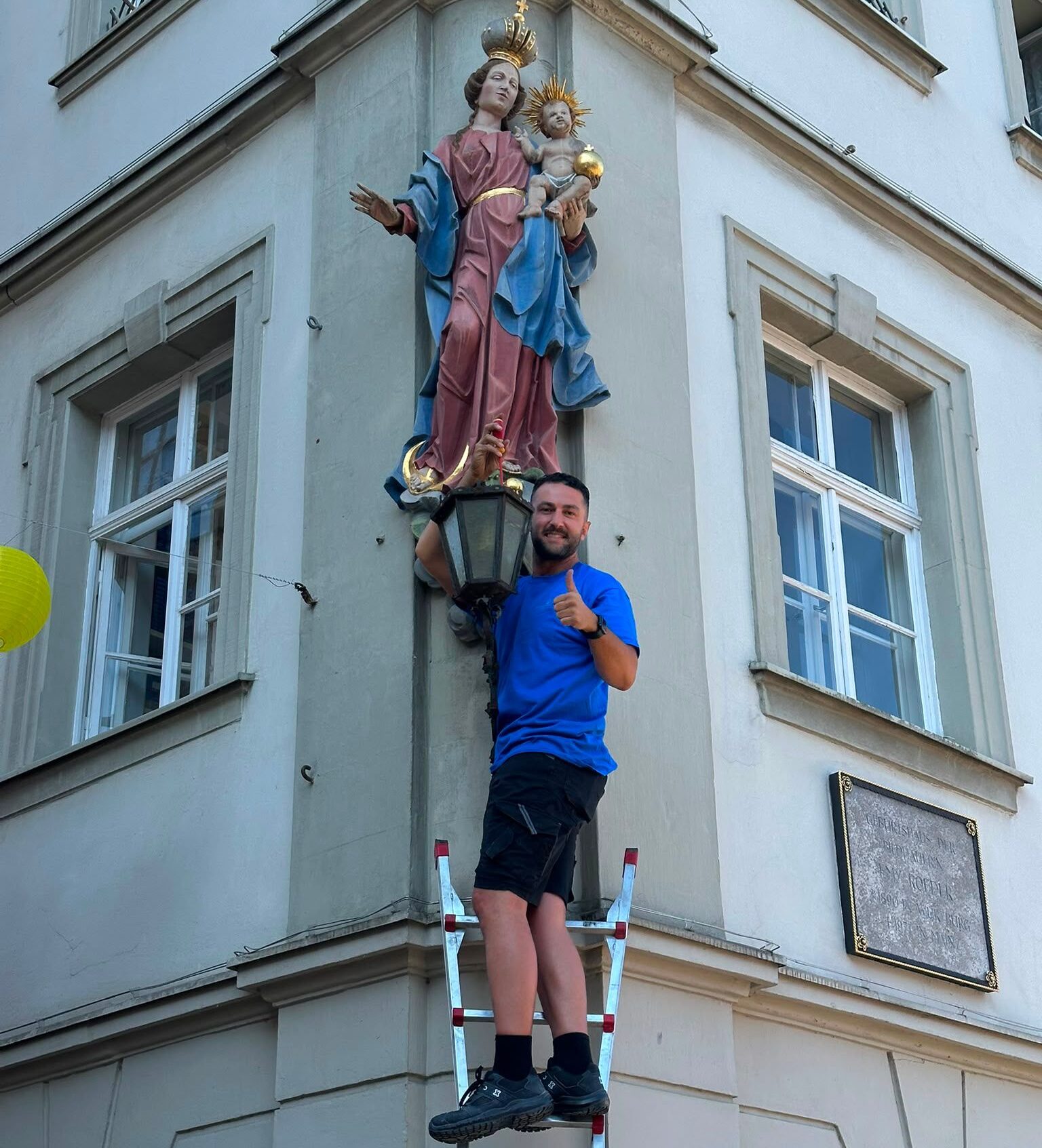 Elektromeister Kemal Marasli mit Daumen hoch auf der Leiter vor einer Hausfassade in der Würzburger Altstadt